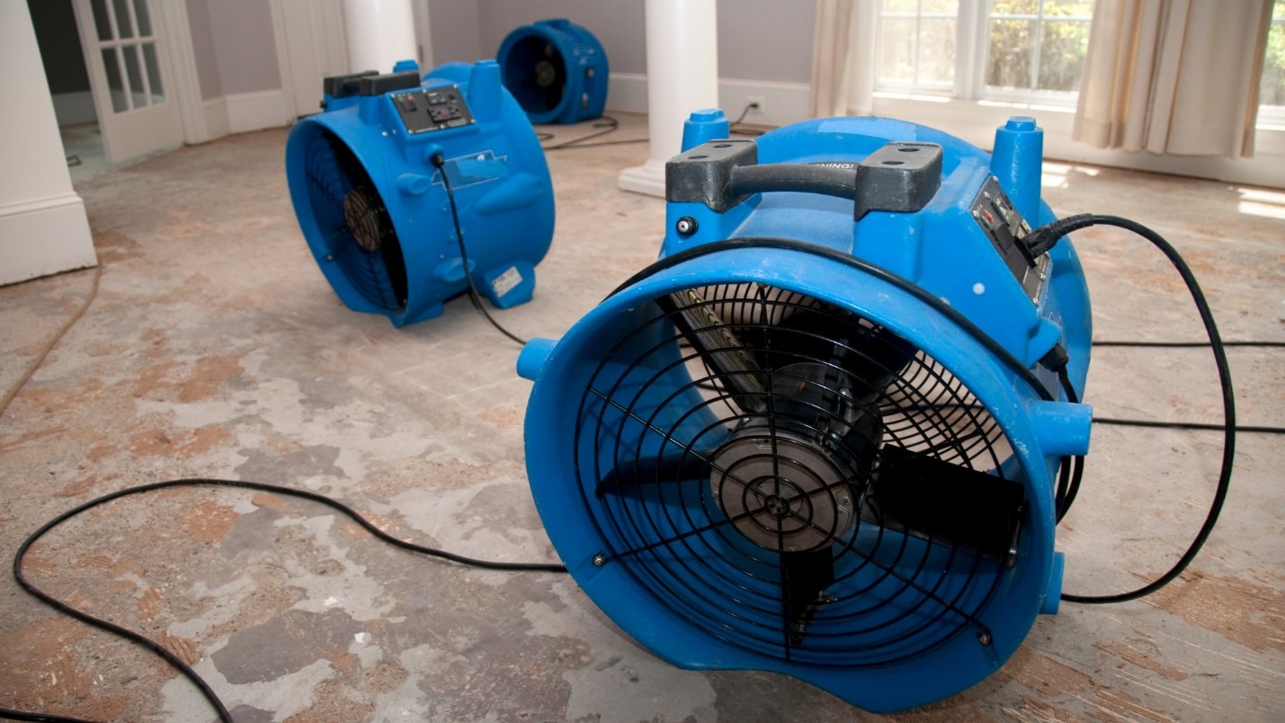 Three large blue industrial grade fans sit on a water damaged floor.